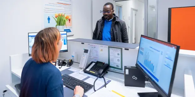Mann steht am Empfang der Präventionsambulanz, spricht mit einer Mitarbeiterin am Schreibtisch mit zwei Monitoren.